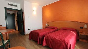 A hotel room with two single beds, a wooden headboard, a bedside table with a lamp, a small table with a chair, and a door leading to another room.