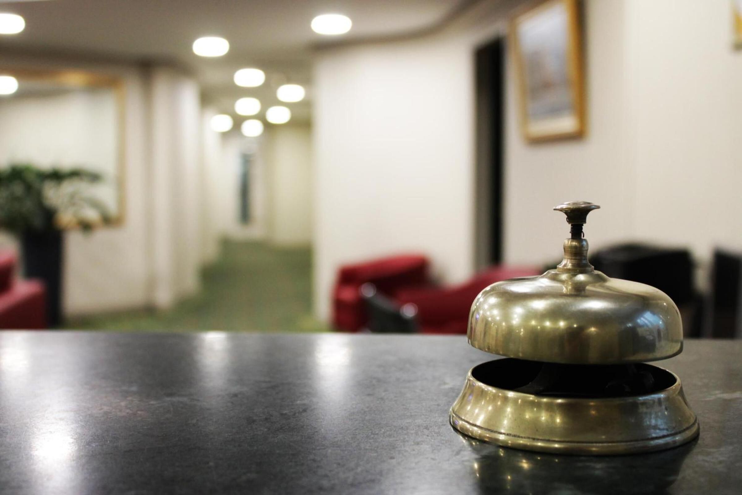 A hotel reception area with a metallic bell on a counter, a red chair, and a plant in the background.