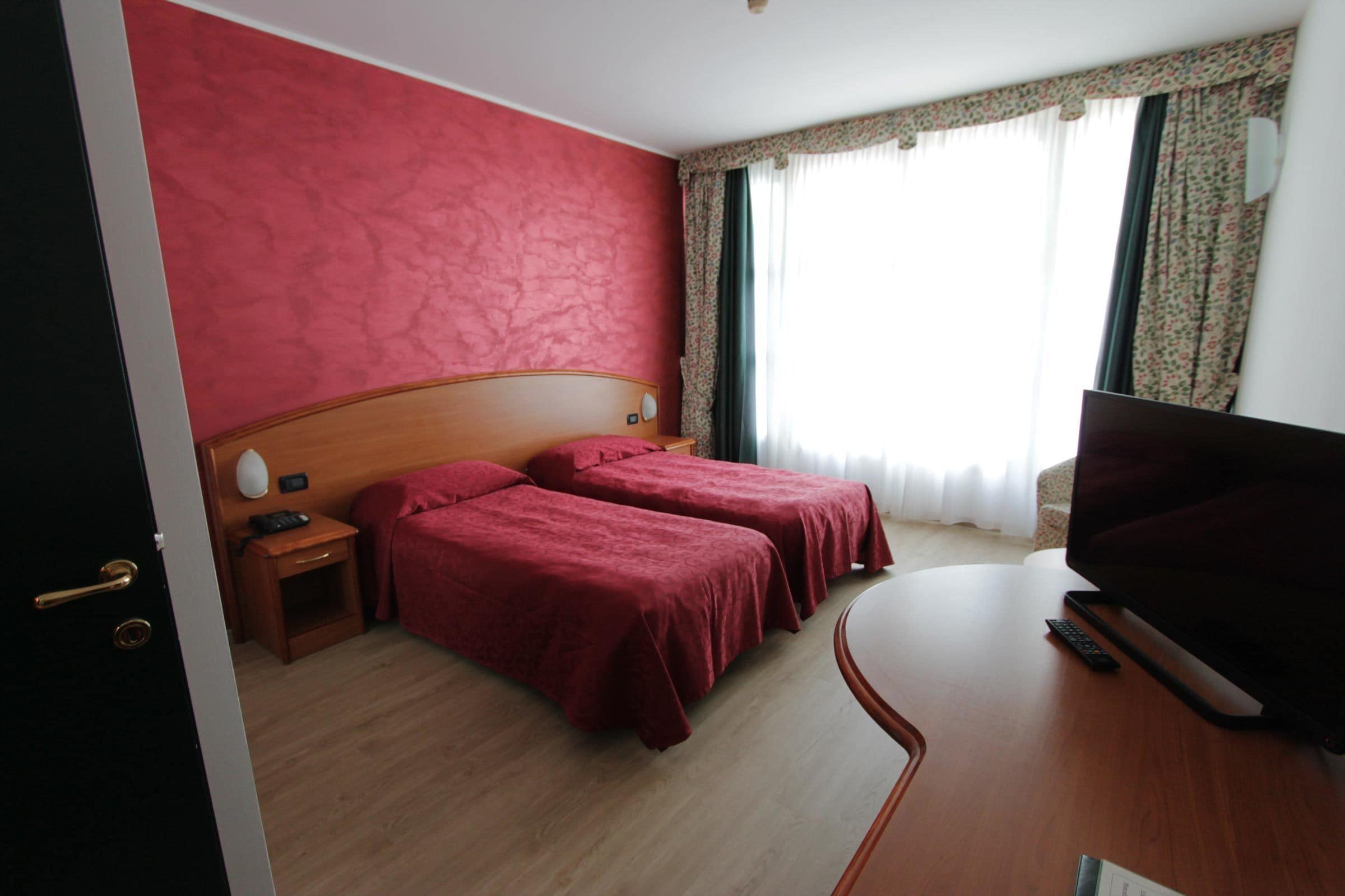 A hotel room with two single beds, a wooden headboard, a flat-screen TV, a bedside table with a lamp, and a window with curtains.