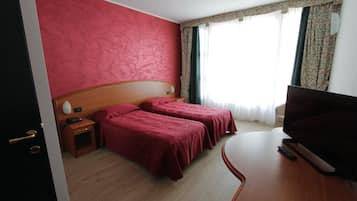 A hotel room with two single beds, a wooden headboard, a flat-screen TV, a bedside table with a lamp, and a window with curtains.