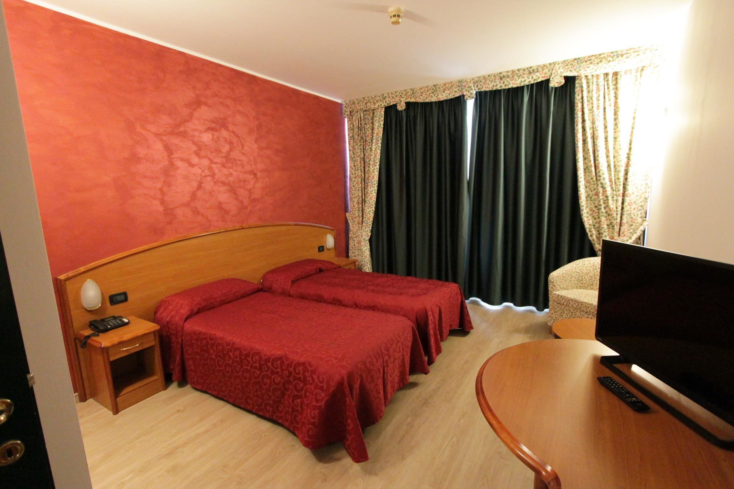 Quarto de hotel com duas camas de solteiro, cabeceira de madeira, mesa de cabeceira com abajur, televisão e uma cadeira.