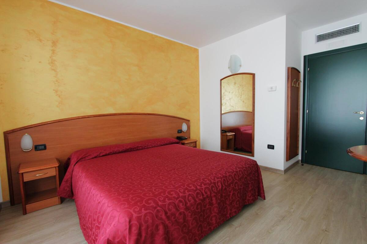 Ein Hotelzimmer mit einem hölzernen Kopfteil, einer rot gemusterten Bettdecke, einem Nachttisch mit Lampe, einem Spiegel und einer grünen Tür.