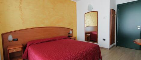 Una habitación de hotel con cabecera de madera, colcha estampada de color rojo, mesita de noche con lámpara, espejo y puerta verde.