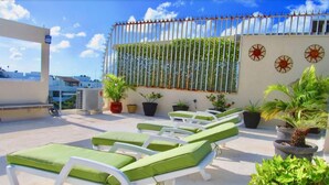 Een terras op het dak met witte lounge fauteuils en groene kussens, omringd door potplanten en decoratieve zonmotieven op de muur.
