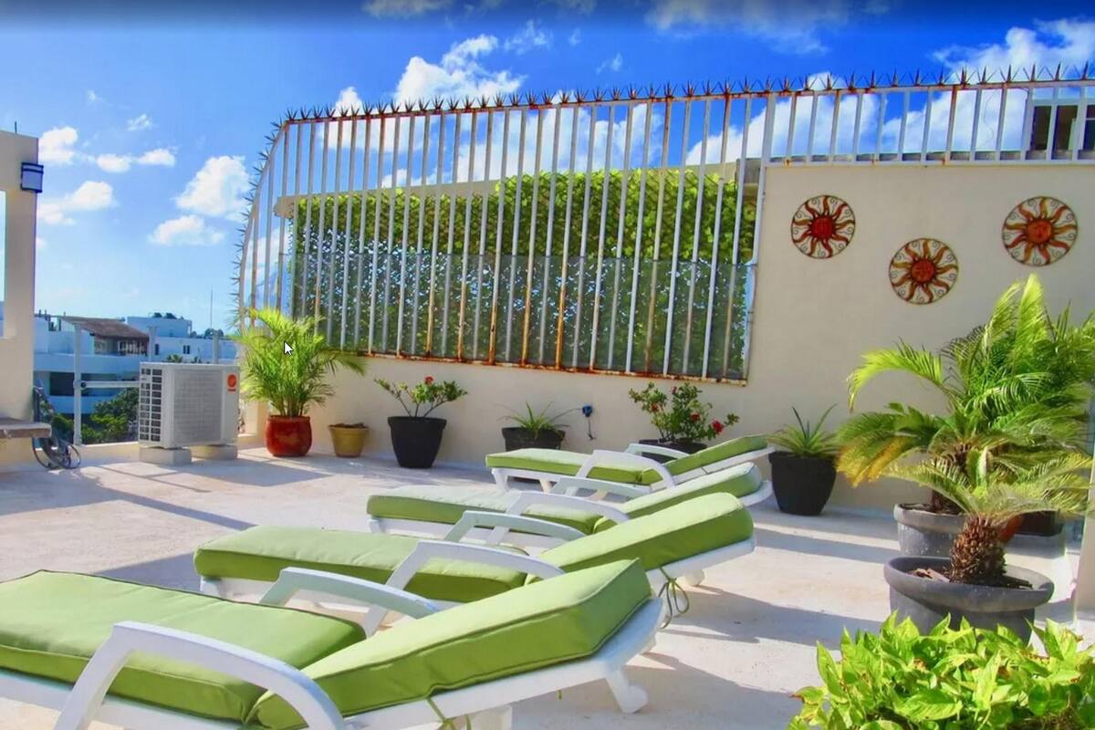 Una terraza en la azotea con sillones blancos y cojines verdes, rodeada de plantas en macetas y motivos solares decorativos en la pared.