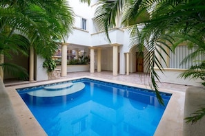 A swimming pool with a tiled deck, surrounded by palm trees and a white building with columns.