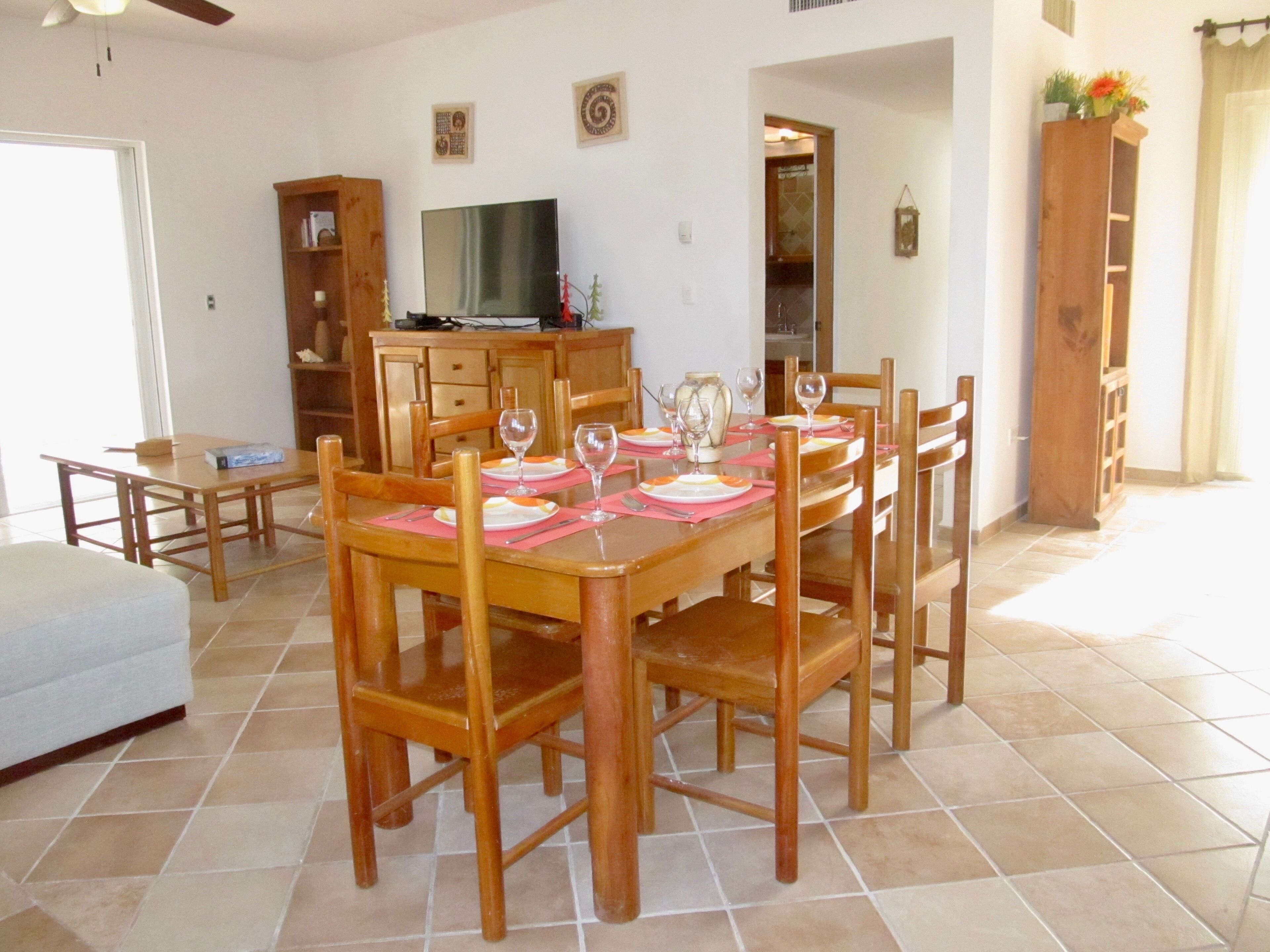 A dining area with a wooden table set for four, surrounded by chairs, with a television on a wooden cabinet in the background.