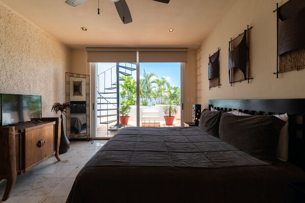 A bedroom with a large bed, a TV on a wooden cabinet, a potted plant, and a balcony with plants and a staircase. - Bosque de los Aluxes Condos with private pool by PPR (Playa del Carmen)