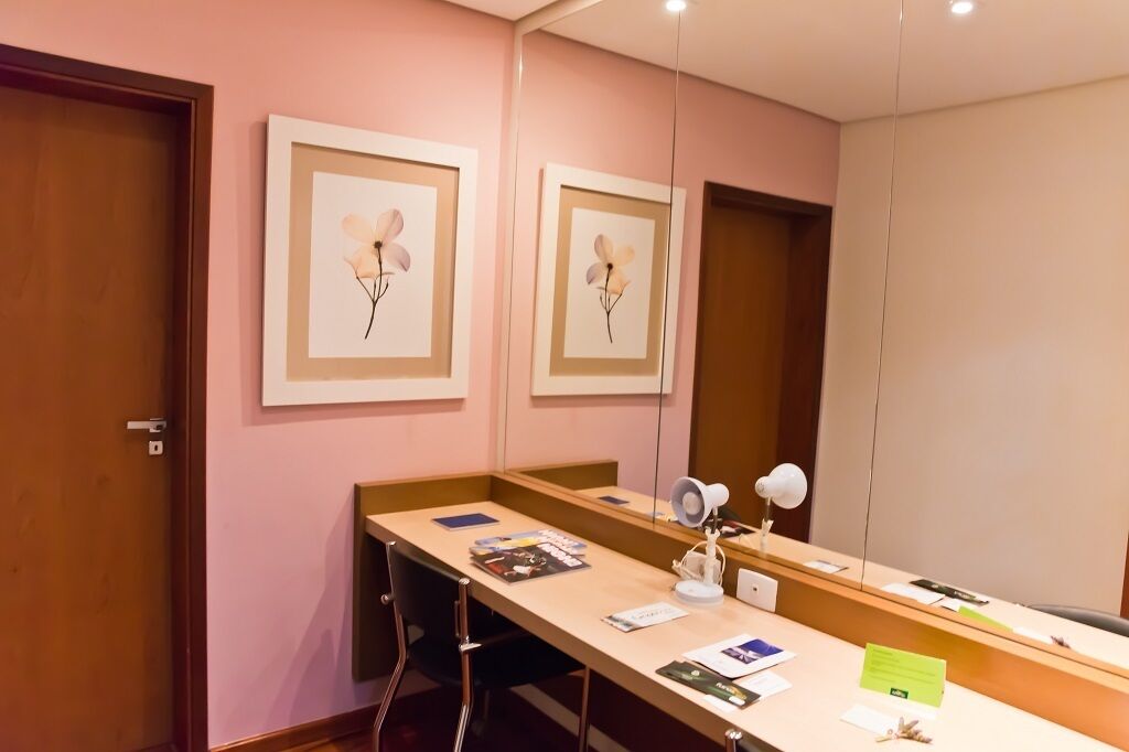 A hotel room with a wooden desk, a chair, a mirror, and framed floral artwork on the wall.