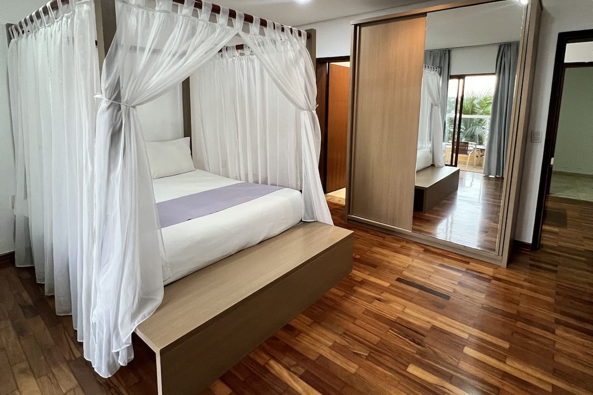 Un dormitorio moderno con cama con dosel, suelo de madera y un espejo grande que refleja la habitación.