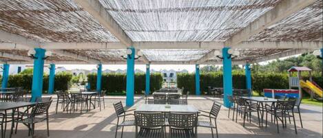 An outdoor patio area with tables and chairs, covered by a woven ceiling, and surrounded by blue pillars.