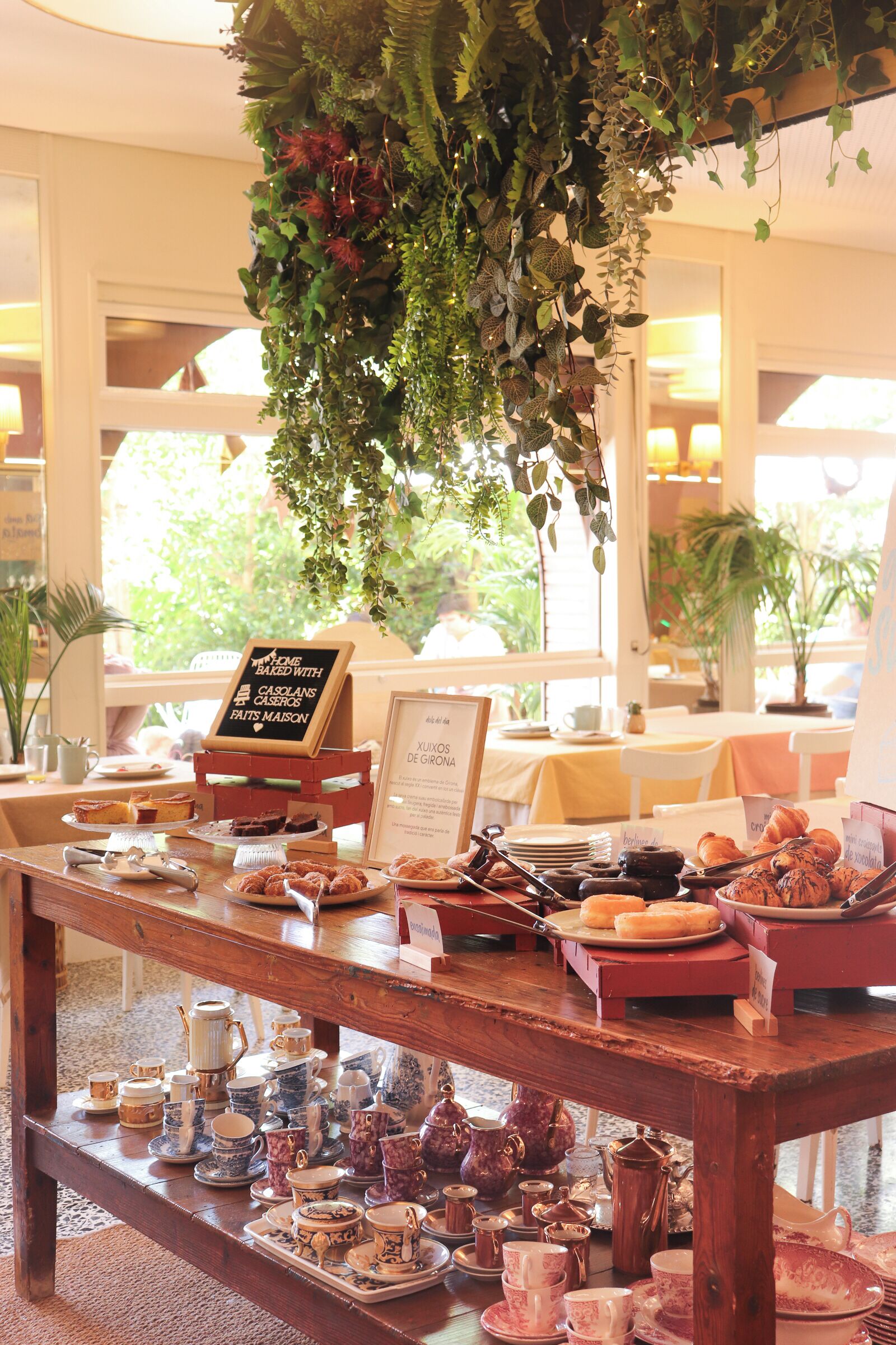 Un café aménagé avec une variété de produits de boulangerie et de pâtisseries présentés sur une table en bois, un menu écrit à la craie et des plantes suspendues au-dessus.