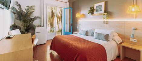 A hotel room with a bed, bedside tables, a desk, a chair, a potted plant, and a view of palm trees through the door.