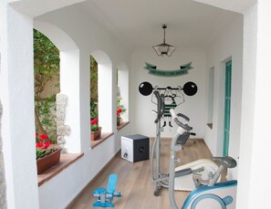 Un'area esterna coperta con attrezzature per l'esercizio fisico, tra cui una cyclette e un vogatore, e una parete con una citazione motivazionale.