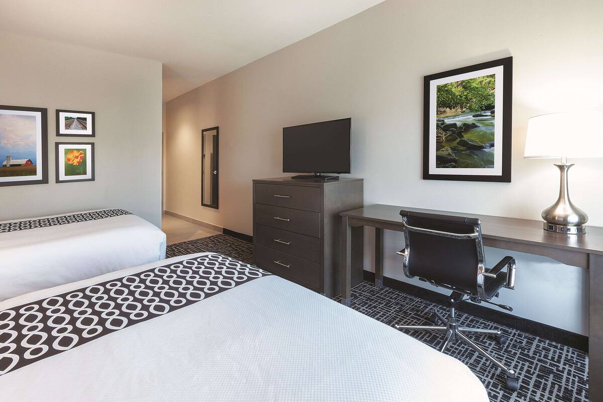 A hotel room with two beds, a desk with a chair, a TV, and framed pictures on the wall.