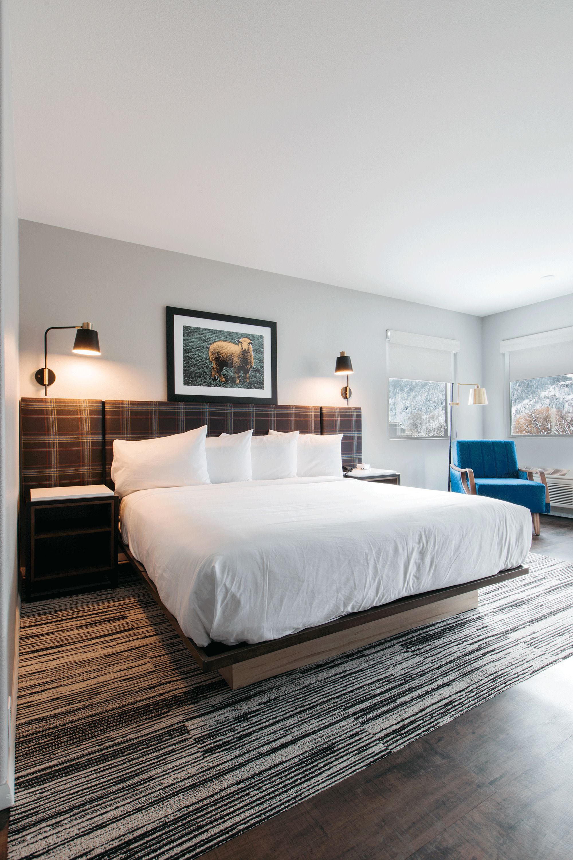 A modern hotel room with a large bed, a blue armchair, and a framed picture on the wall.