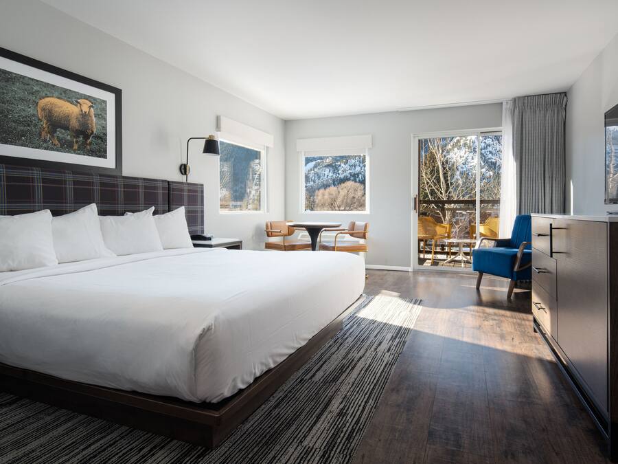 A modern hotel room with a large bed, a dining area, and a view of the outdoors.