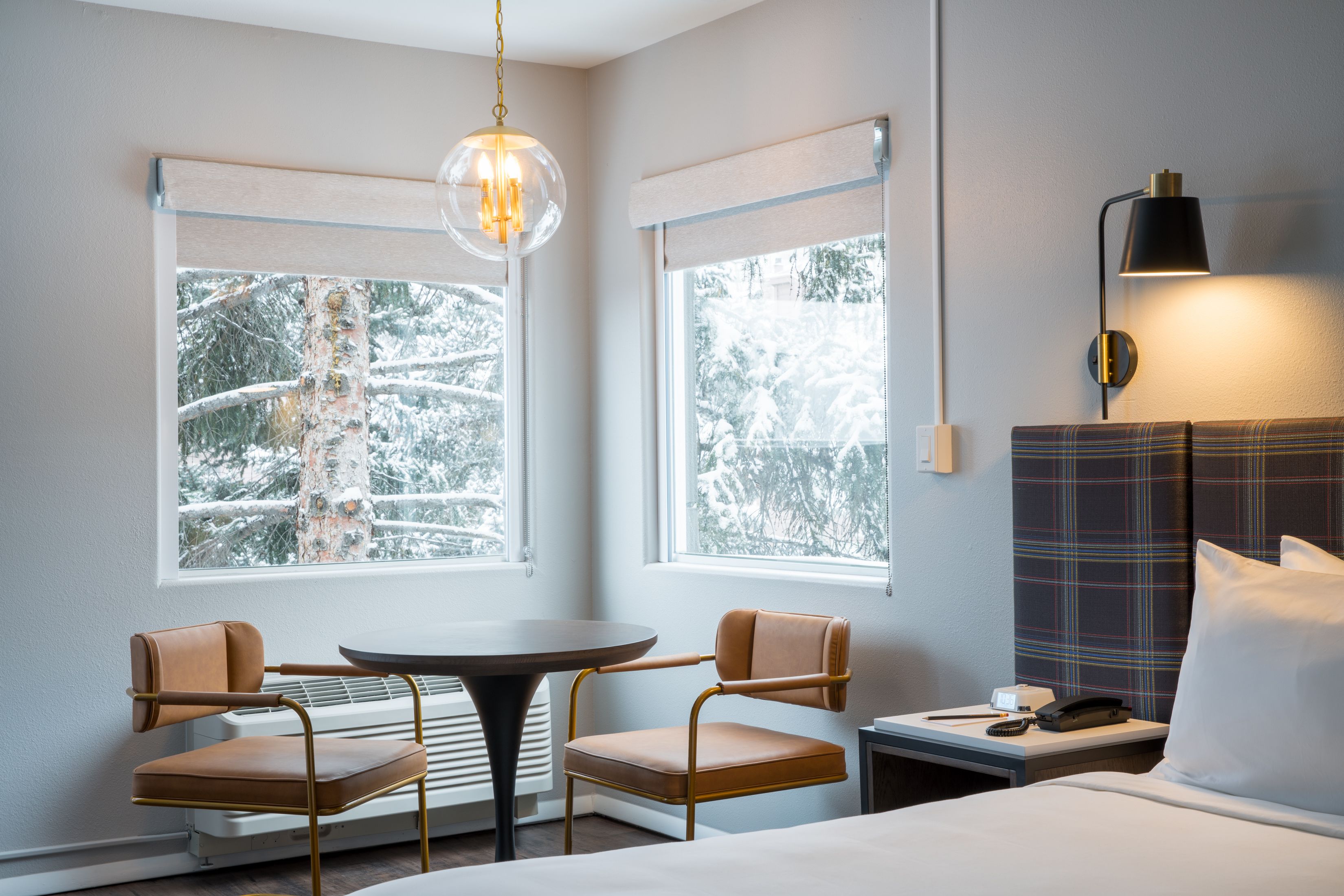 Quarto de hotel com uma cama, uma mesa redonda, duas cadeiras, mesa de cabeceira com abajur e janelas com vista para árvores cobertas de neve.