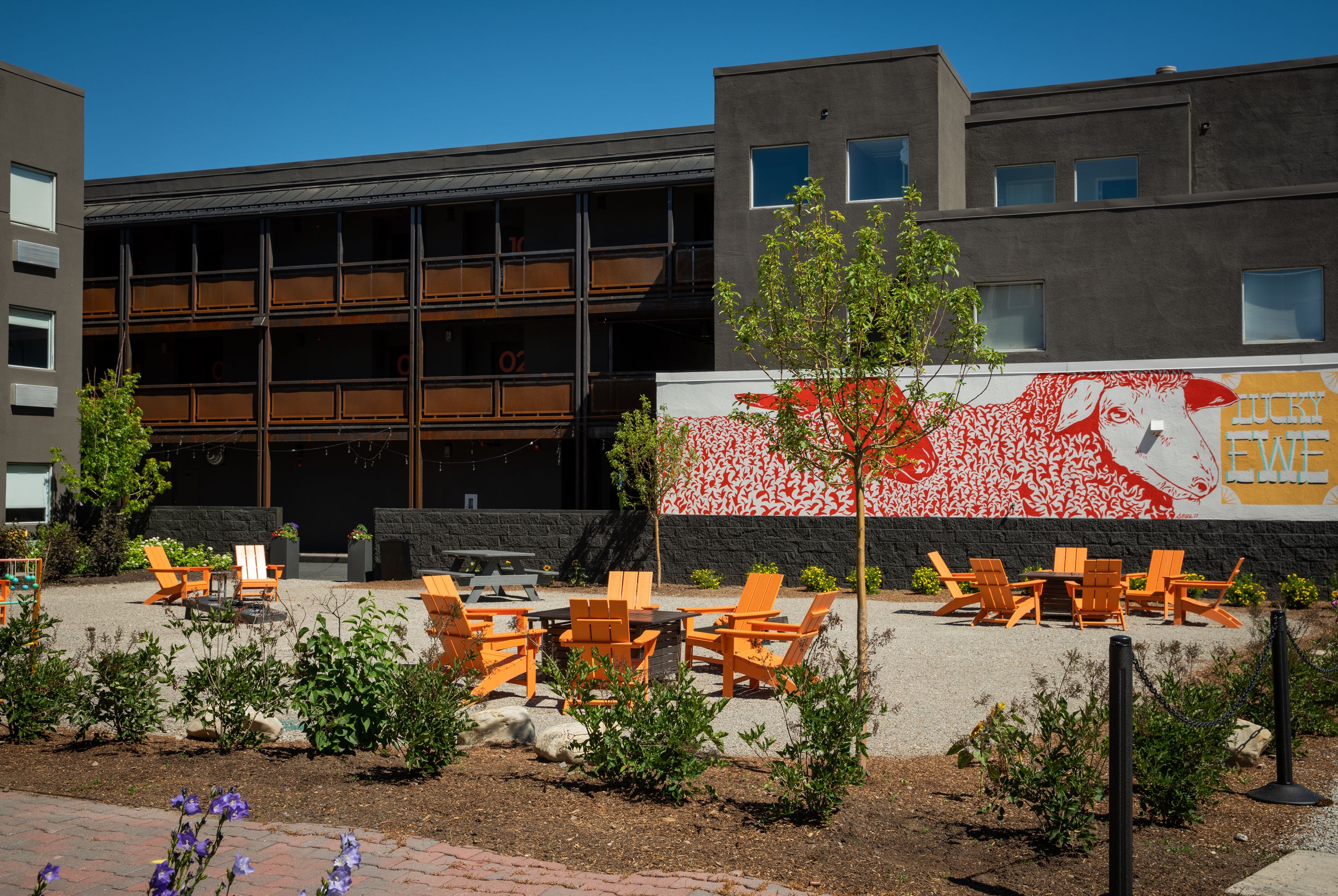 Un espace extérieur aménagé avec des chaises orange et une table, un bâtiment orné d’une grande fresque murale, et un ciel d’un bleu éclatant.