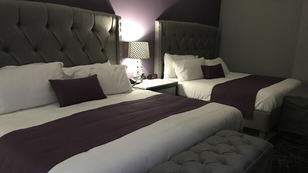 A hotel room with two beds, each with a tufted headboard, white bedding, and purple accents. There is a small table with a lamp between the beds.
