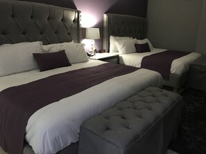 A hotel room with two beds, each with a tufted headboard, white bedding, and purple accents. There is a small table with a lamp between the beds.