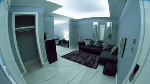 A modern living room with a sofa, a coffee table, and a TV. There is a desk with a computer and a chair. The room has a large window and a ceiling with a skylight.