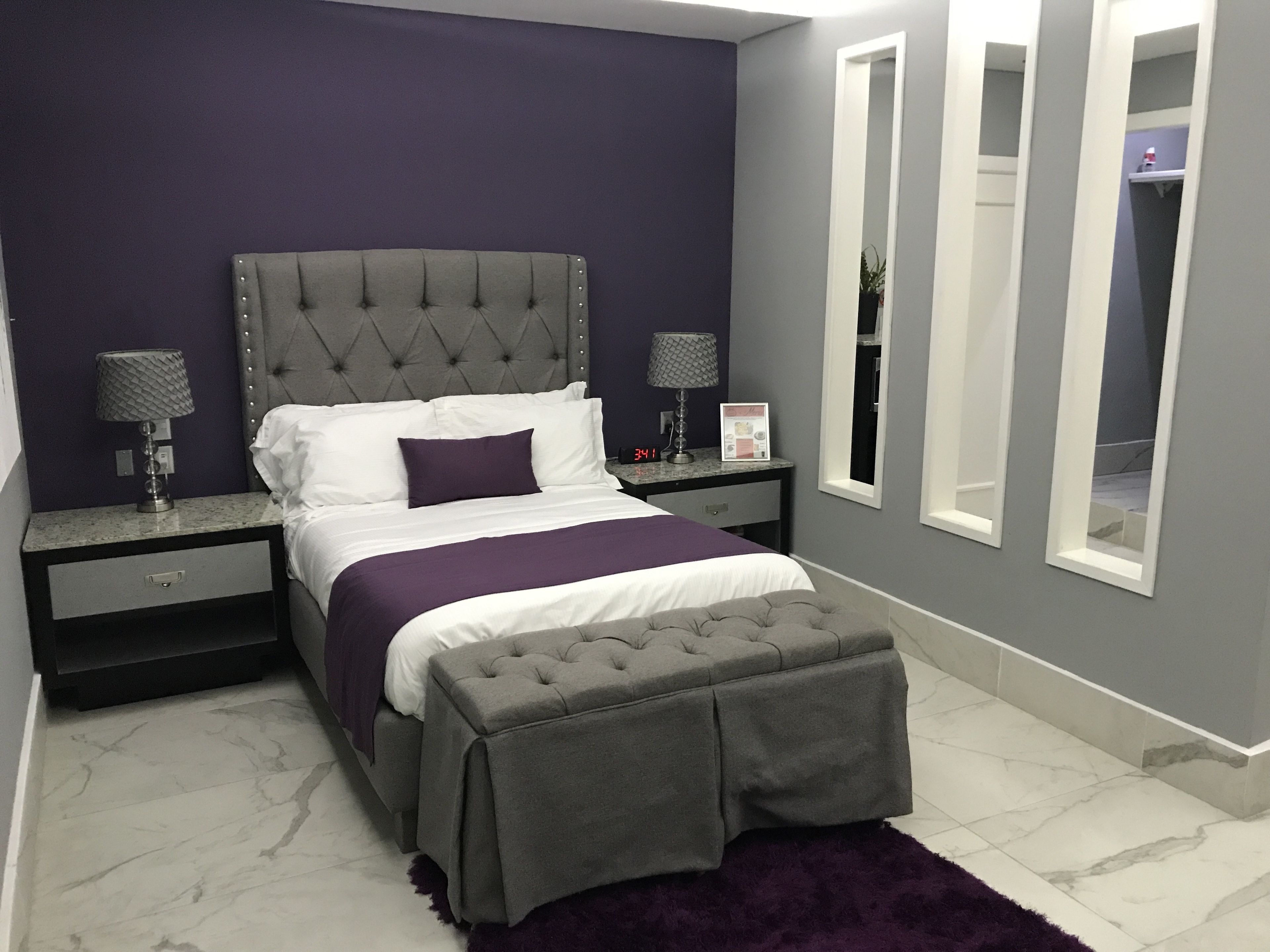 Un dormitorio moderno con un cabecero capitonado, un banco gris y una manta morada.