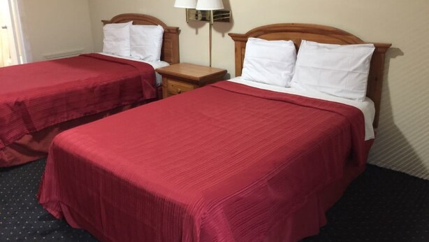 A hotel room with two beds, each with a red bedspread and white pillows. There is a nightstand with a lamp between the beds. - Royal Inn (Honea Path)