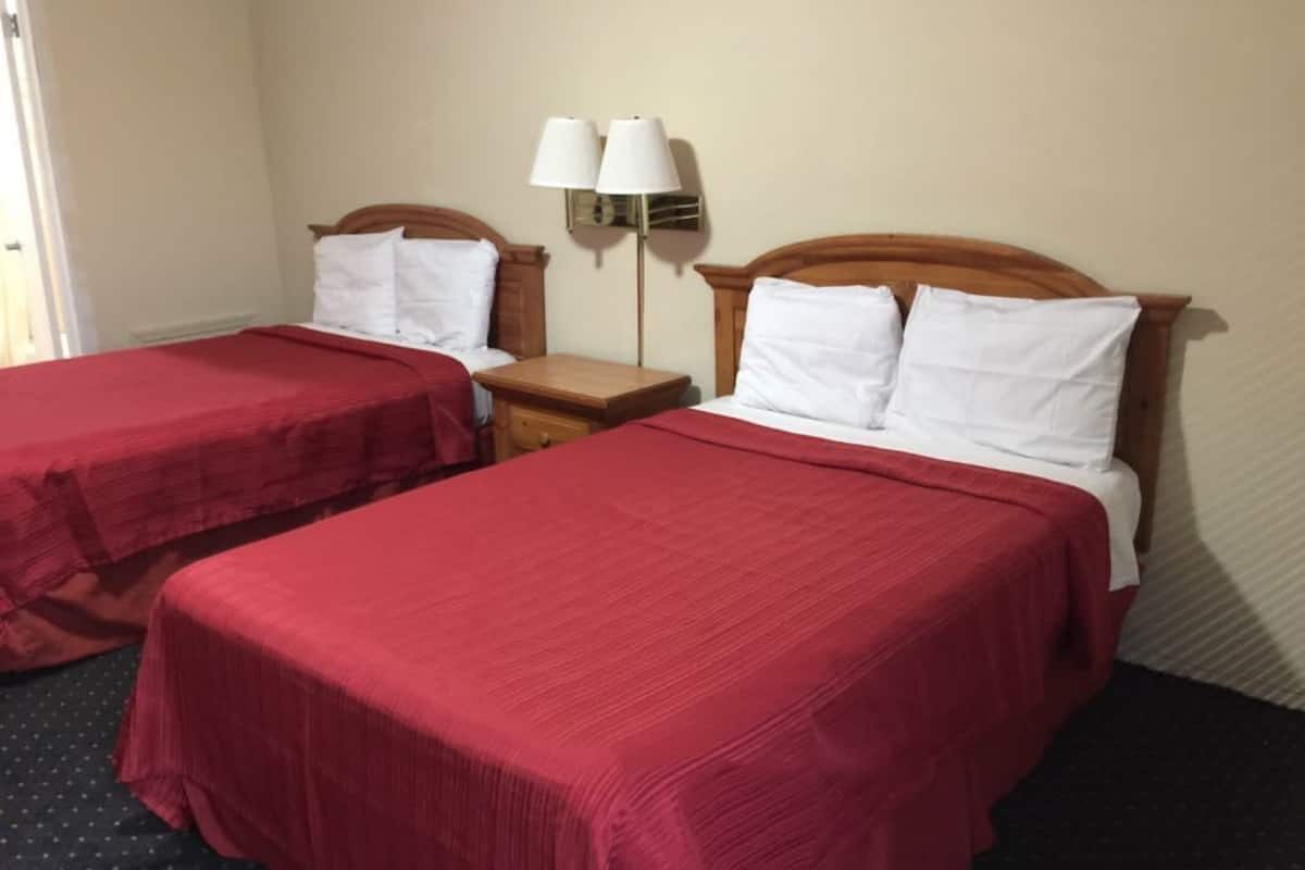 A hotel room with two beds, each with a red bedspread and white pillows. There is a nightstand with a lamp between the beds.