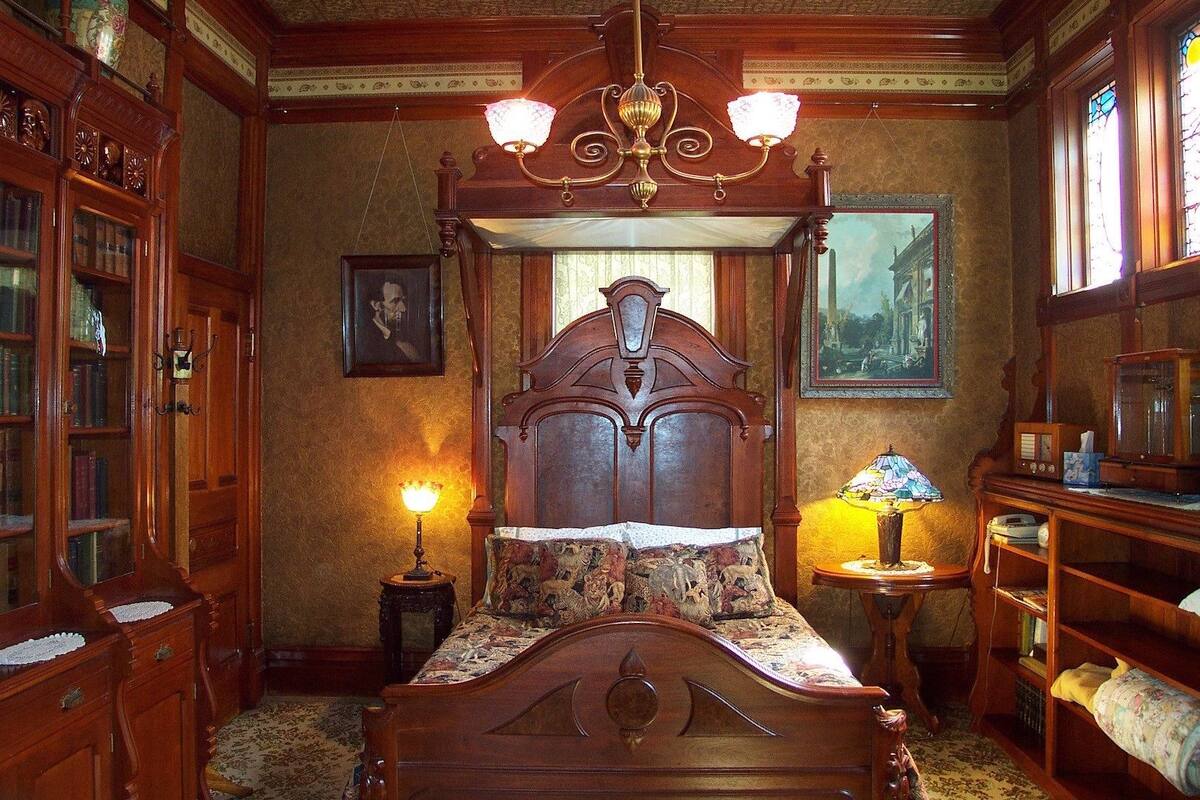 Un lit à baldaquin avec une couvre-lit à motifs floraux, un lustre, une petite table avec une lampe et une bibliothèque remplie de livres.