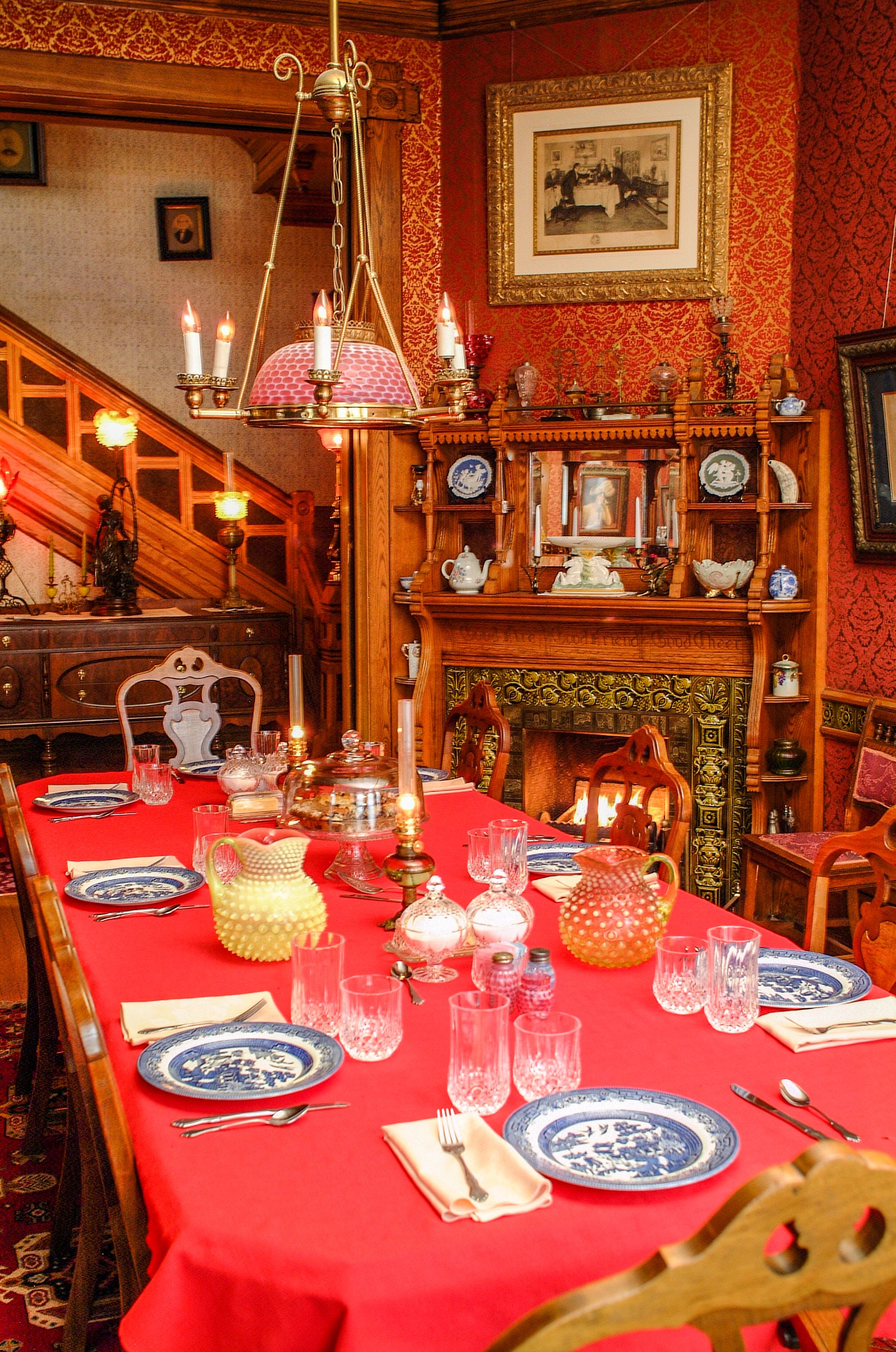 A dining room with a red tablecloth, ornate wooden furniture, and a fireplace.