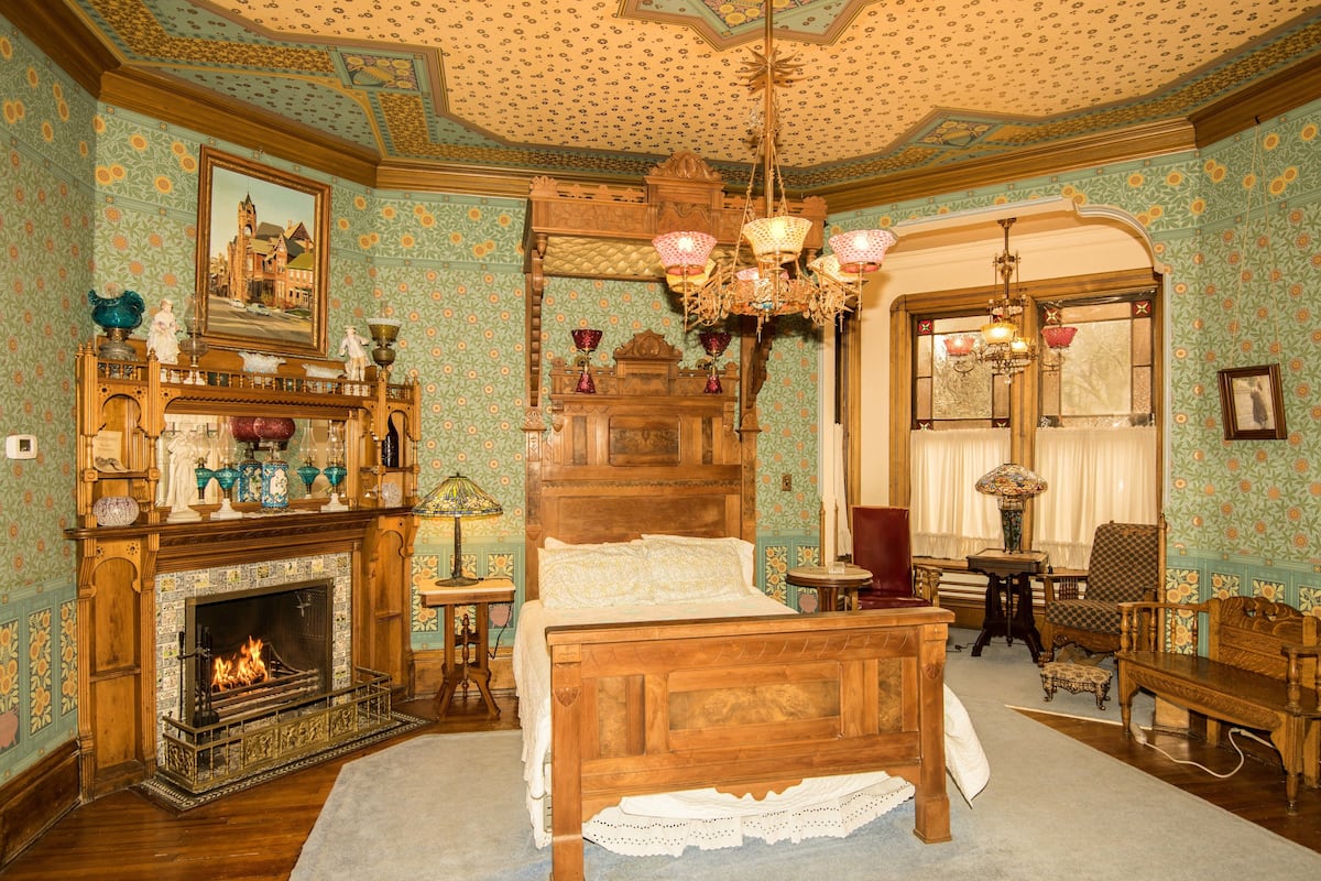 Une pièce avec un lit en bois, une cheminée, un lustre et un papier peint à motifs.