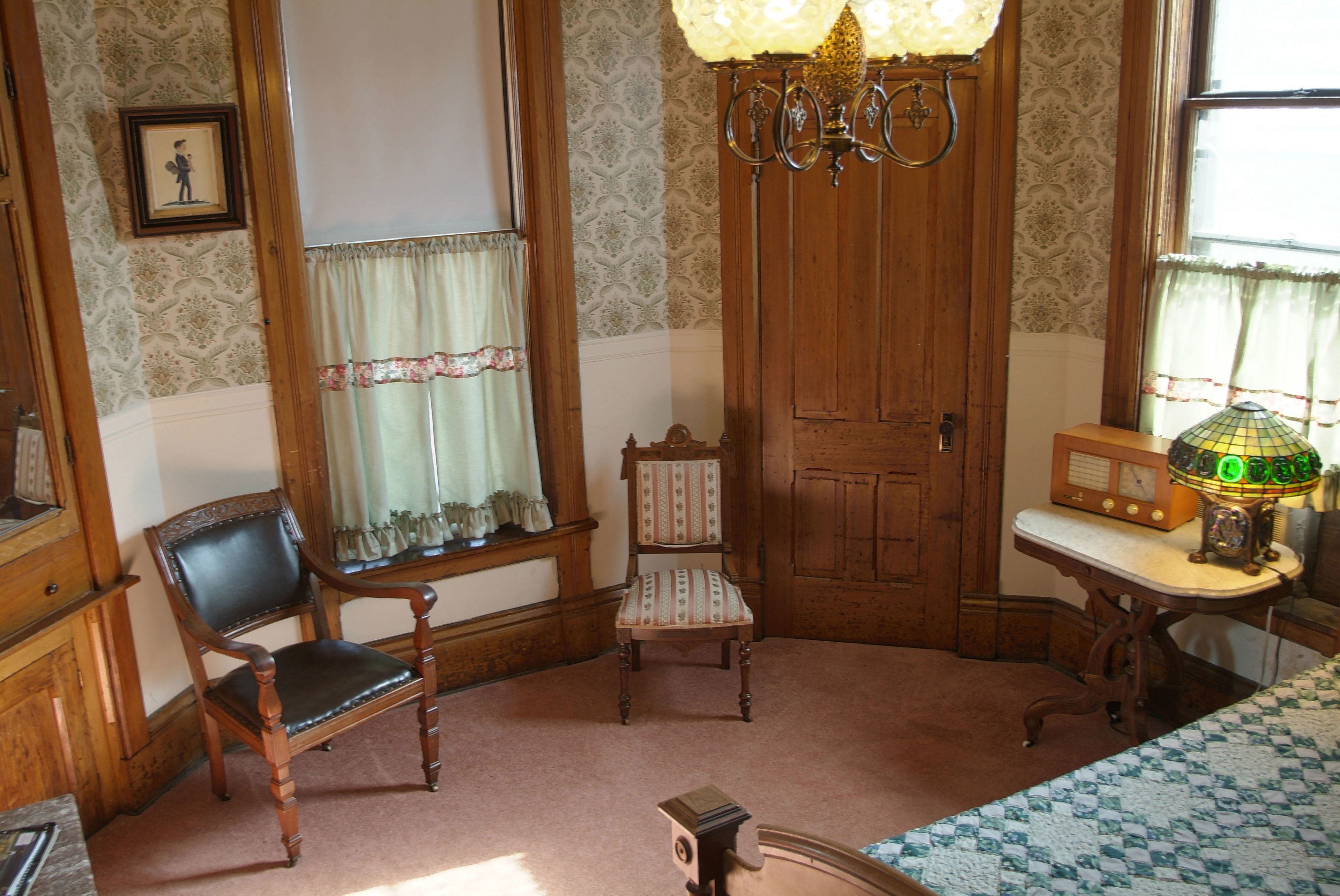 A room with two wooden chairs, a table with a radio, and a window with curtains.