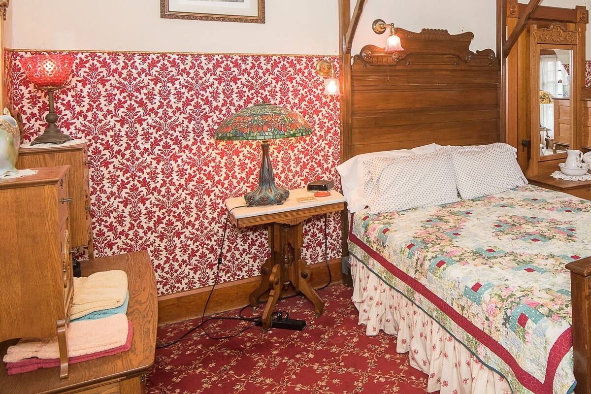 Une chambre à coucher de style traditionnel, avec un lit en bois, un papier peint à motifs floraux, une commode, une chaise et une lampe.