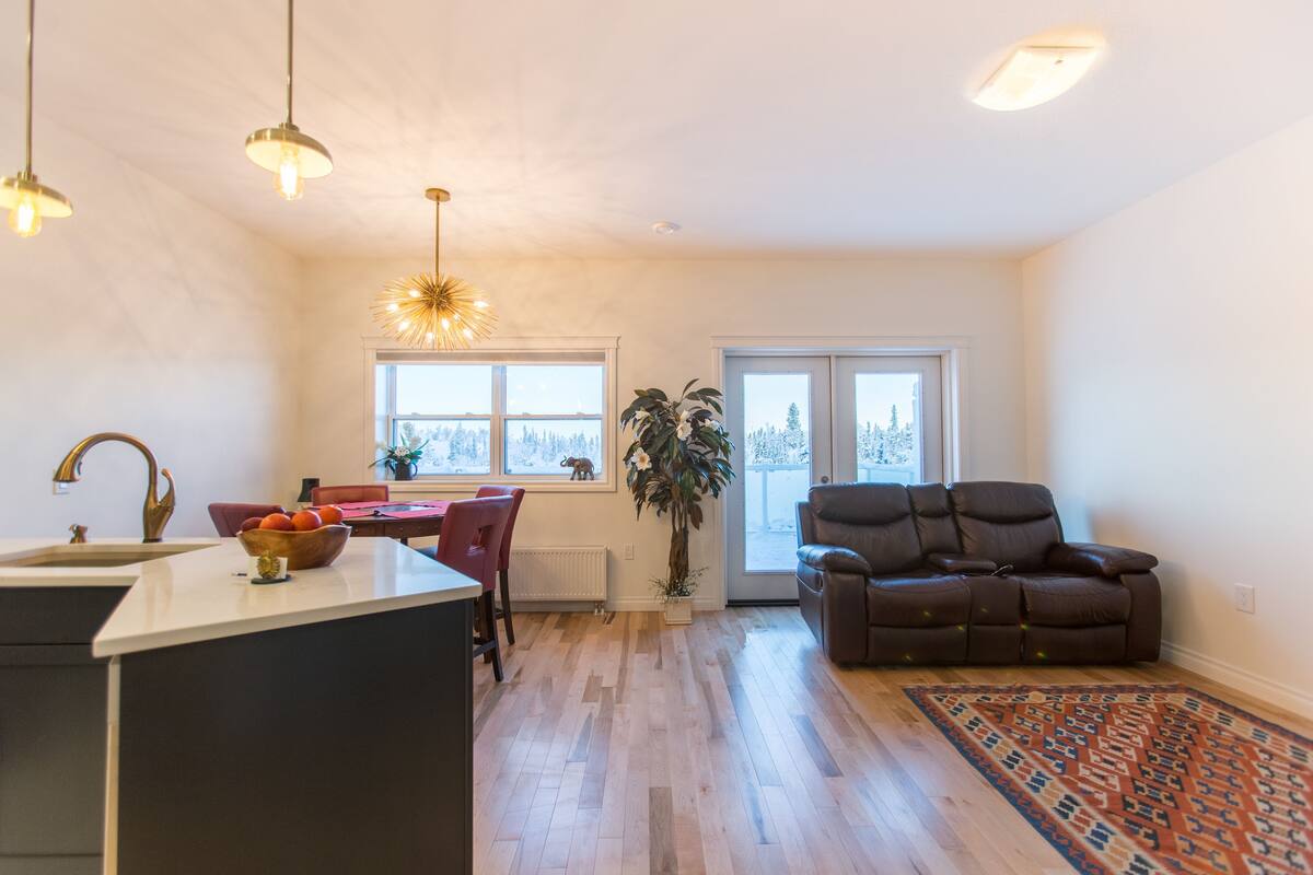 A modern kitchen with a central island, a dining area with a red chair, a brown leather sofa, and a patterned rug.