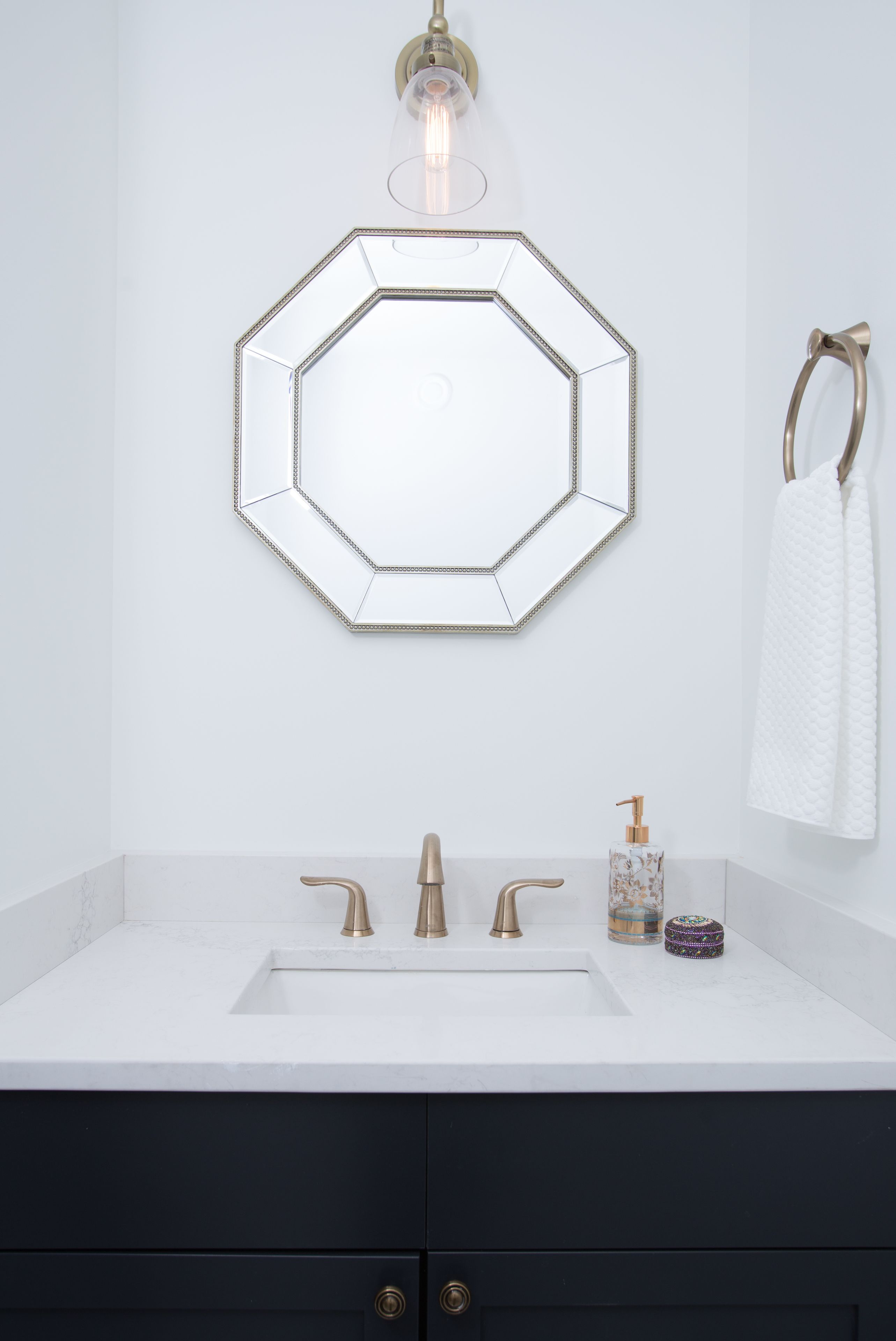Ein Badezimmer mit einem weißen Waschbecken, drei goldenen Wasserhähnen, einem sechseckigen Spiegel, einem Handtuchhalter und einer Seifenspender.