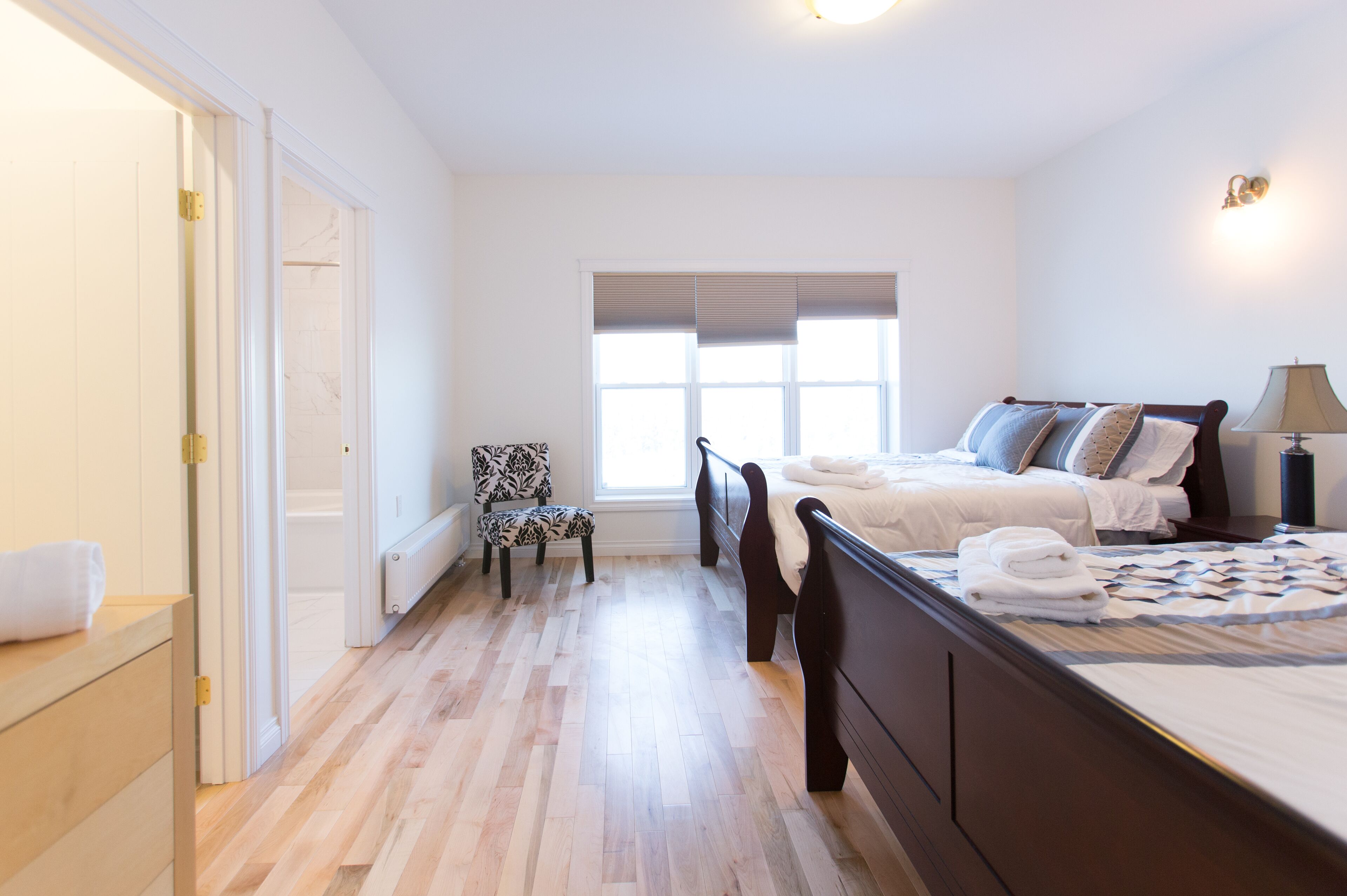 A bedroom with a large bed, a chair, a window with blinds, and a bedside table with a lamp.
