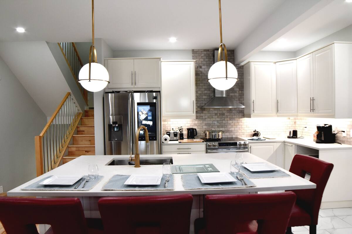 A modern kitchen with white cabinets, a stainless steel refrigerator, a built-in oven, and a dining area with red chairs and a white table set for four.
