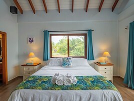 Habitación de hotel con una cama grande, dos mesitas de noche con lámparas, una ventana con cortinas y una colcha con motivos tropicales.