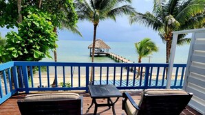 A wooden deck with a table and chairs, overlooking a beach with a thatched structure and palm trees.