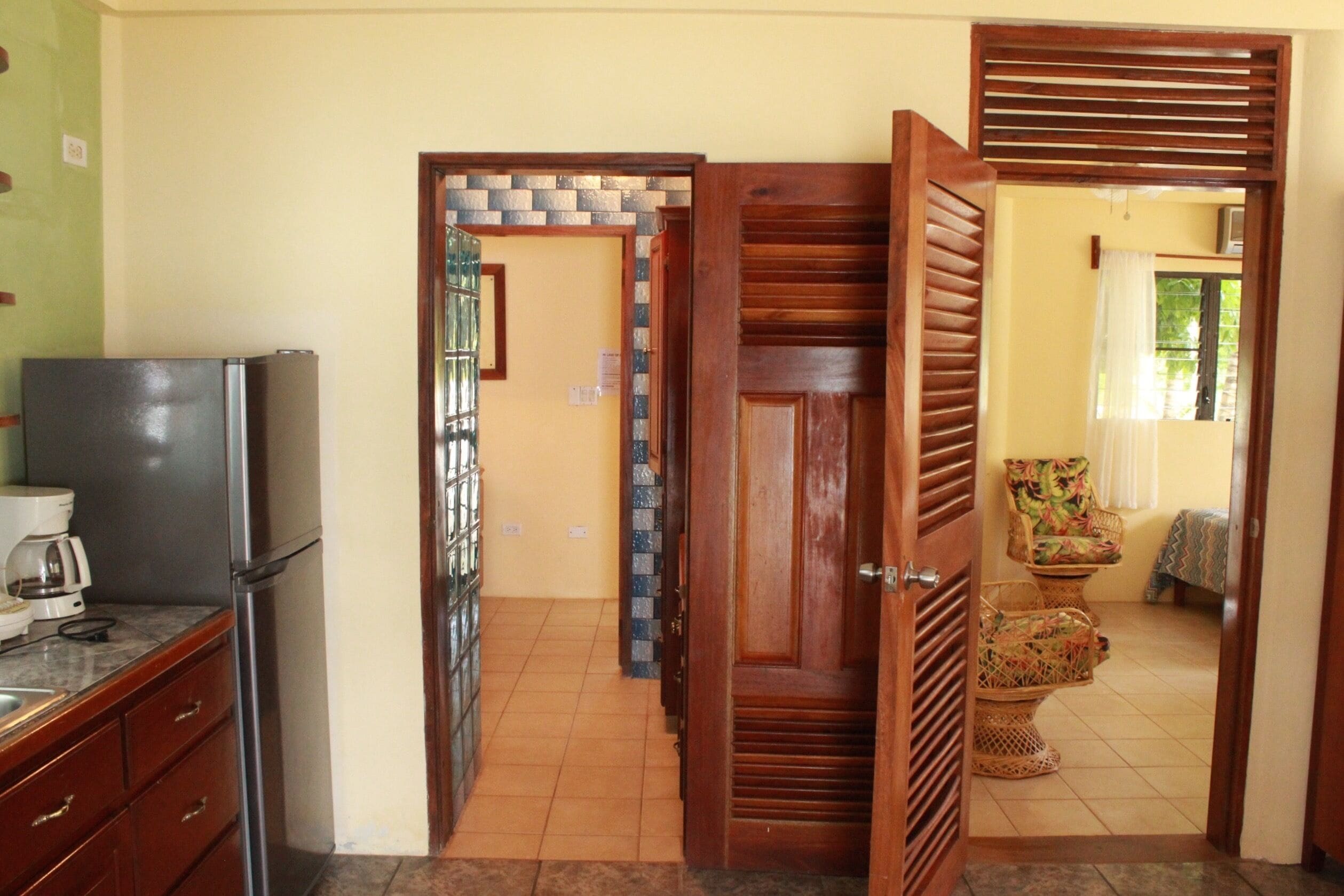 A kitchen with a refrigerator, a coffee maker, and a tiled floor. There is a wooden door leading to another room with a chair and a bed.