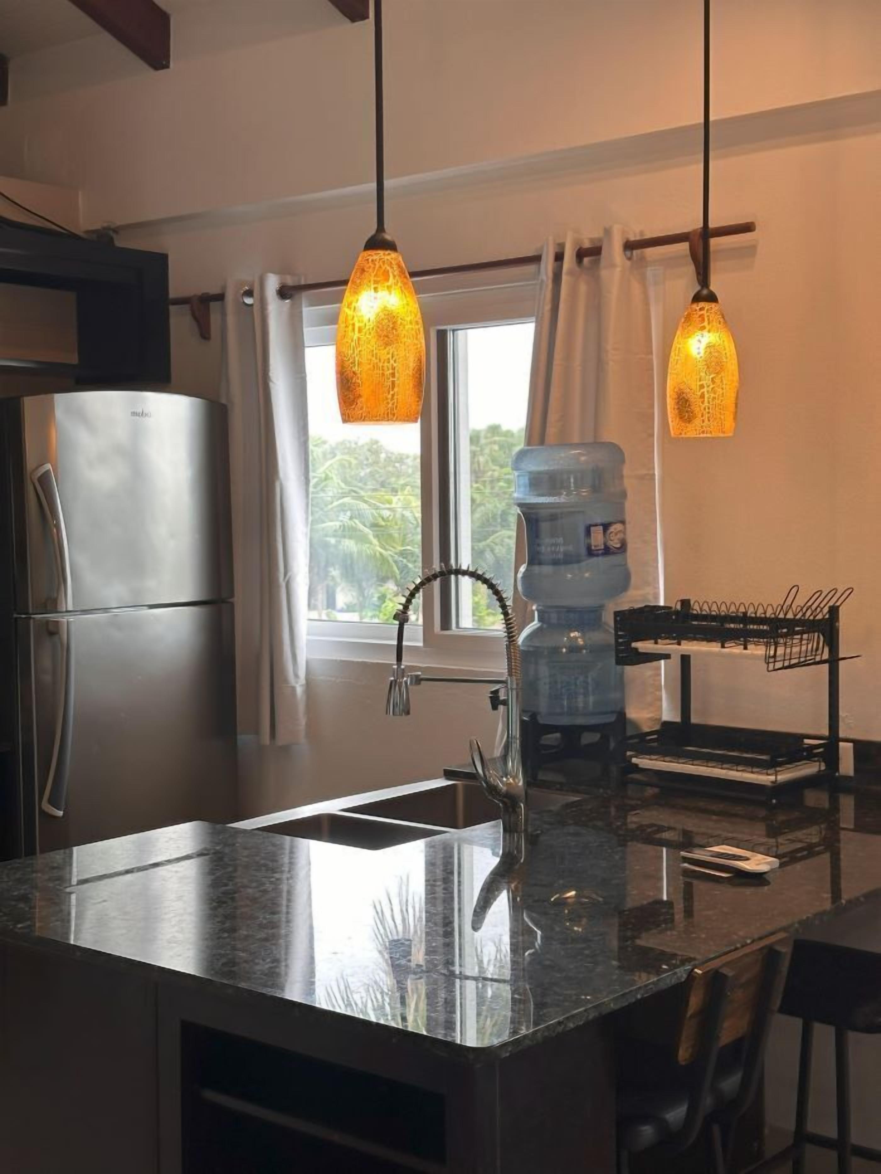 A modern kitchen with a stainless steel refrigerator, a sink with a unique faucet, and pendant lights.