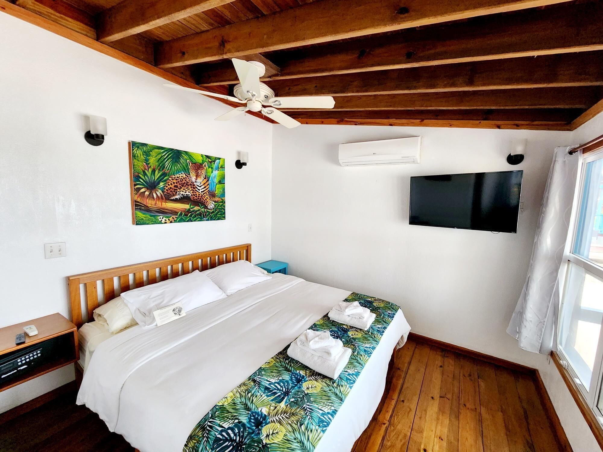 A hotel room with a wooden ceiling, a bed with a tropical print bedspread, a mounted TV, and an air conditioning unit.
