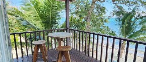 A wooden deck with two stools and a table, overlooking a beach with palm trees and the ocean.