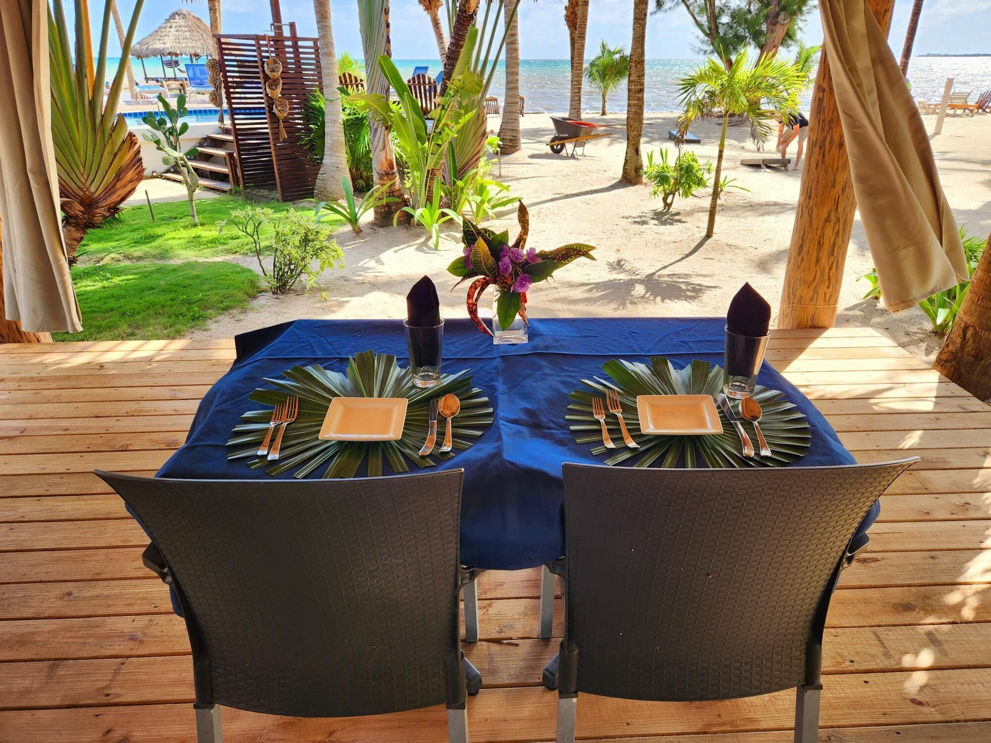A wooden deck with a table set for two, overlooking a beach with palm trees and a clear view of the ocean.