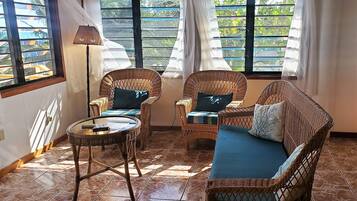 Una habitación con mobiliario de mimbre, incluyendo un sofá y dos sillones, una mesa redonda y ventanas grandes con cortinas.