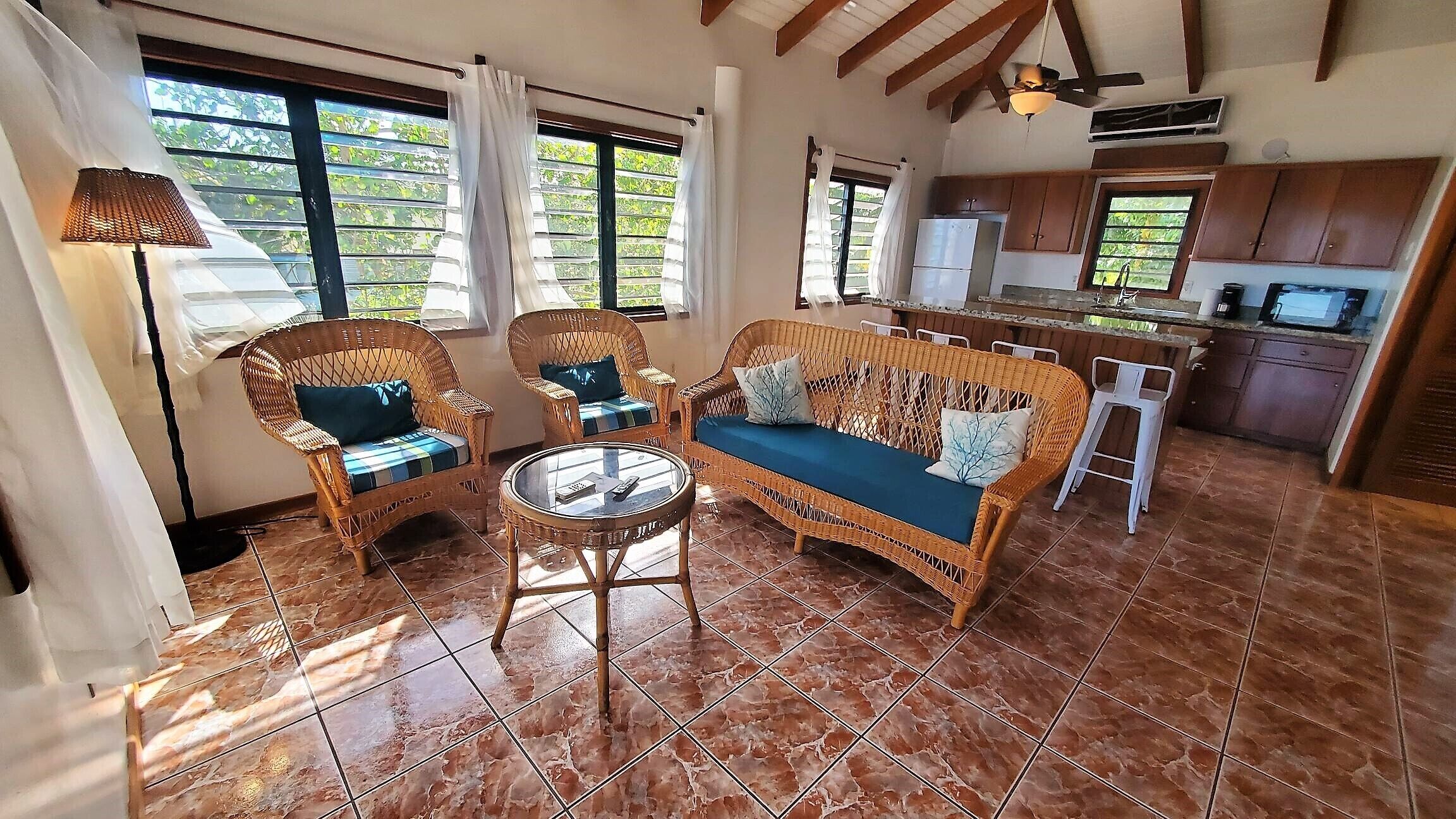 Un espacio de sala acogedor con muebles de mimbre, una mesa de vidrio y una cocina al fondo.