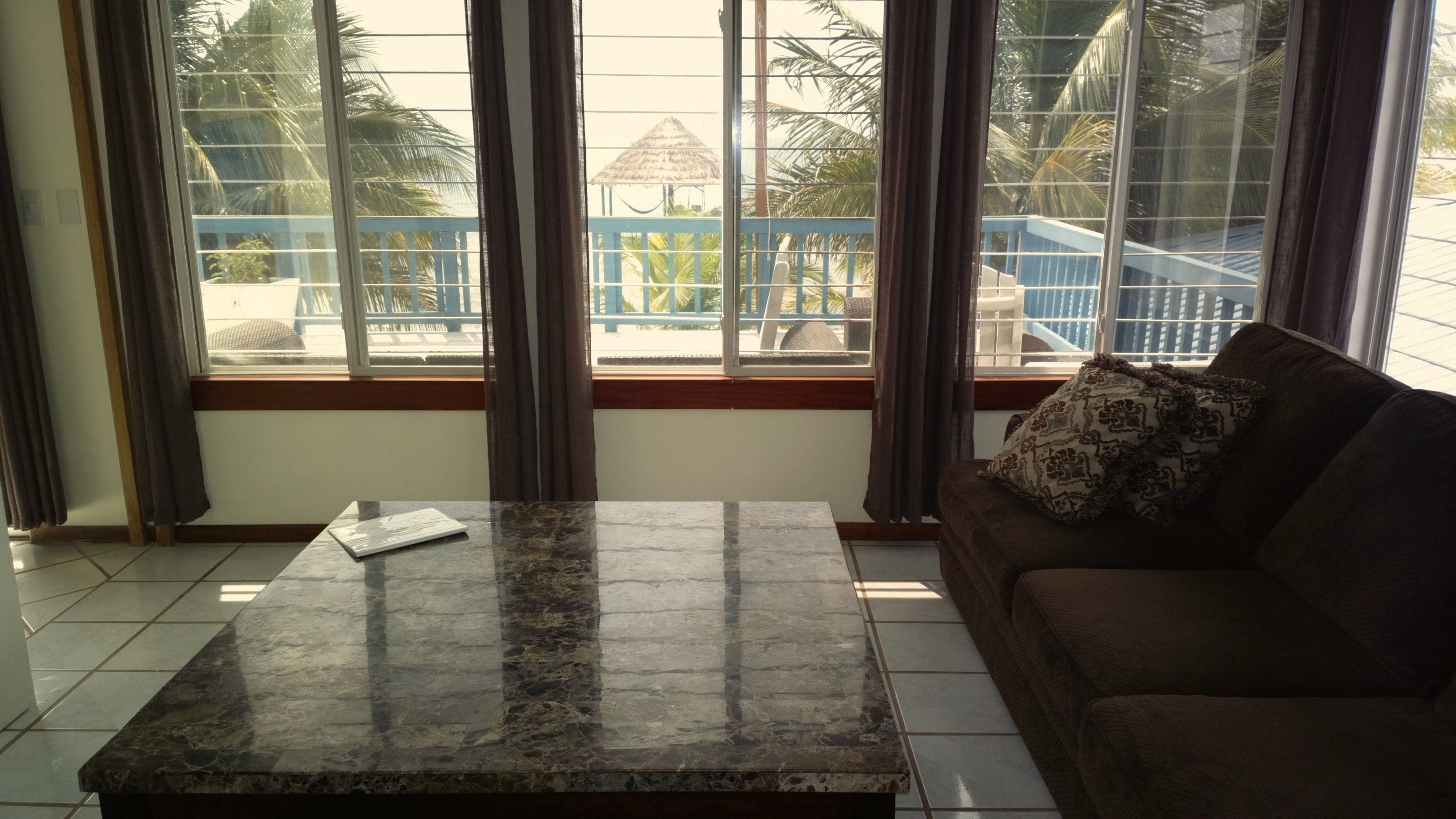 A room with a marble coffee table, a brown sofa with a patterned pillow, and large windows with a view of a pool and palm trees.
