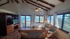 A room with a ceiling fan, a dining table with chairs, a bookshelf, and large windows with a view of the ocean.