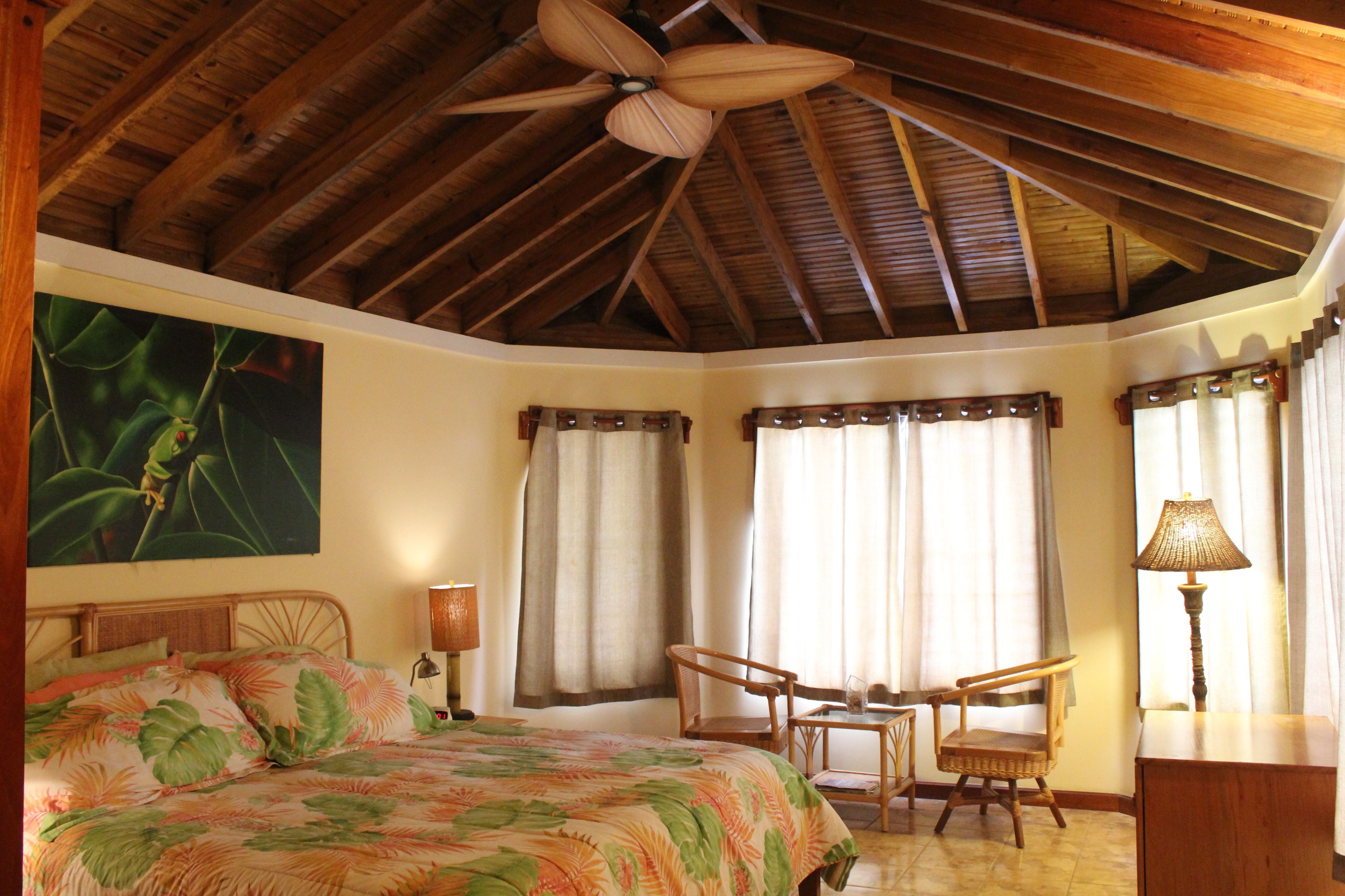 A bedroom with a bed, two chairs, a desk, a ceiling fan, and a painting on the wall.